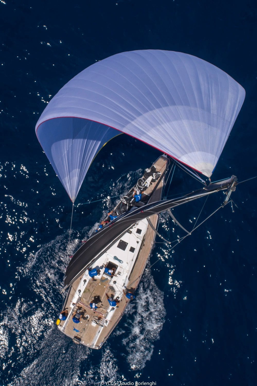 Aerial view of a racing sailboat under spinnaker, crew on board in full action.